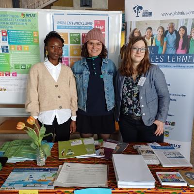 © WUS 2025 Drei junge Frauen stehen lächelnd hinter einem Infostand zum Thema Globales lernen.