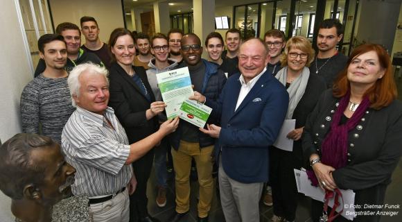 Auf diesem Bild sieht man eine Gruppe von Menschen, die sich zur Zertifikatsverleihung versammelt haben. Im Vordergrund überreichen zwei Personen ein Zertifikat: ein Mann mit grauem Haar und eine Frau in schwarzem Blazer, während sie von weiteren Teilnehmern begleitet werden. Die anderen Personen in der Gruppe, die im Hintergrund stehen, schauen aufmerksam zu. Das Bild wurde anscheinend bei einer feierlichen Veranstaltung aufgenommen, die von mehreren Personen aus verschiedenen Bereichen besucht wurde.