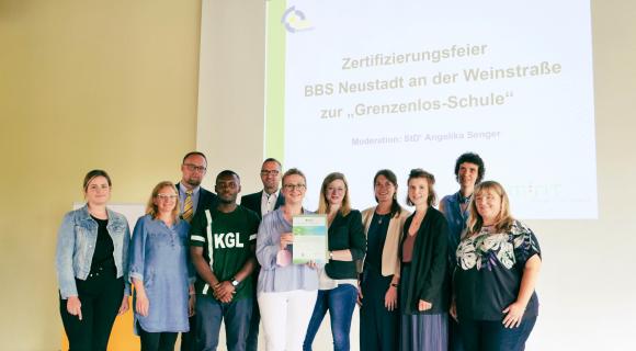 Eine Gruppe von 13 Personen steht vor einer Präsentationsfolie mit der Aufschrift „Zertifizierungsfeier BBS Neustadt an der Weinstraße zur ‚Grenzenlos-Schule‘“. Zwei Personen in der Mitte halten ein Zertifikat. Die Gruppe wirkt stolz und lächelt in die Kamera. Der Raum hat eine helle, freundliche Atmosphäre, und alle tragen formelle oder legere Kleidung. Die Stimmung ist feierlich und engagiert.