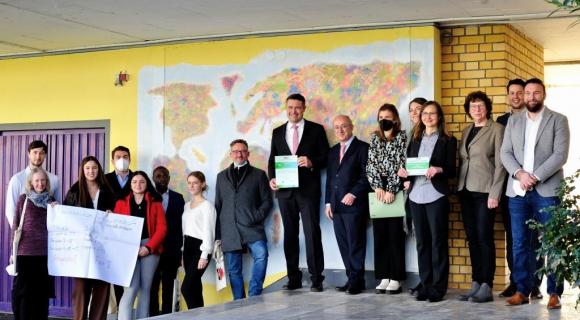 Das Bild zeigt eine Gruppe von Menschen, die vor einer Wand mit einer Weltkarte stehen. Einige Personen halten Urkunden in den Händen, andere stehen nebeneinander. Die Gruppe besteht aus Erwachsenen in formeller Kleidung sowie einigen jüngeren Menschen. Im Vordergrund ist auch ein großes Plakat zu sehen. Die Stimmung wirkt festlich und feierlich, was auf eine Auszeichnung oder Zeremonie hinweist. Der Raum ist gut beleuchtet und modern eingerichtet.