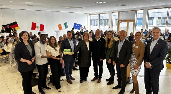 (von rechts: Landrat Olaf Löwis of Menar, Stellvertretende Schulleiterin Susanne Karrer, Ministerialbeauftragter Dr. Marko Hunger, Lehrerin Verena Mayr, Erster Bürgermeister Christoph Schmid, Schulleiterin Cornelia Taube, Ministerialrat Sven Meyer-Huppmann, Grenzenlos-Aktive Barbara Béltran Torres und Ebrottié N’zoo, WUS-Referentin Dr. Julia Boger; FOTO © Peiker 2024 Eine Gruppe von 10 Personen steht zusammen und hält ein Zertifikat in den Händen. Im Hintergrund sind internationale Flaggen zu sehen, darunter Deutschland, Irland und die Europäische Union. Der Raum wirkt festlich und gut besucht, mit weiteren Teilnehmern, die an Tischen sitzen. Die Personen in der Vordergrund strahlen und wirken stolz. Es handelt sich offenbar um eine feierliche Übergabe oder Auszeichnung, die in einer offenen, modernen Umgebung stattfindet.