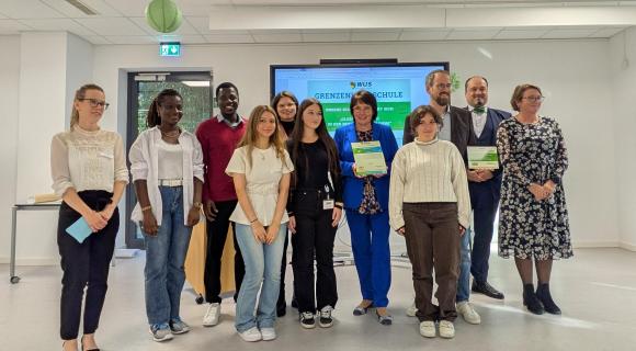 Gruppenbild bei Auszeichnung zur Grenzenlos-Schule ©WUS 2025 Eine Gruppe von Menschen steht in der Aula der Schule. Einige halten Zertifizierungs-urkunden in der Hand.