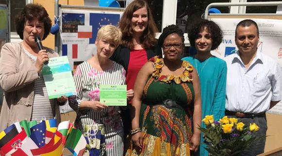 Das OSZ2 in Potsdam erhält Auszeichnung für Globales Lernen im WUS-Projekt „Grenzenlos – Globales Lernen in der beruflichen Bildung Das Bild zeigt eine Gruppe von sechs Personen, die vor einem Gebäude stehen und in die Kamera schauen. Einige halten Zertifikate in den Händen, während andere eine Blume oder ein Mikrofon halten. Im Hintergrund sind Flaggen verschiedener europäischer Länder und Informationsstände zu sehen. Die Personen tragen bunte, sommerliche Kleidung, und die Atmosphäre wirkt festlich und herzlich. Es scheint sich um eine feierliche Veranstaltung oder Auszeichnungszeremonie zu handeln.