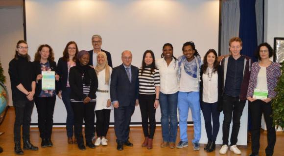 Das Bild zeigt eine Gruppe von 14 Personen, die zusammen auf einer Bühne oder in einem Raum stehen und in die Kamera schauen. Einige halten Zertifikate in den Händen. Die Gruppe besteht aus Menschen unterschiedlichen Geschlechts und unterschiedlicher Herkunft, die in formeller Kleidung oder einem legeren Stil gekleidet sind. Im Hintergrund ist ein heller Raum mit einer Projektionsfläche zu sehen, was auf eine Zeremonie oder ein offizielles Event hindeutet. Alle wirken stolz und engagiert.