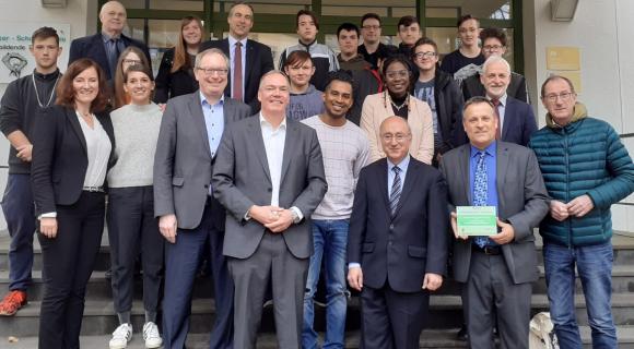Das Bild zeigt eine Gruppe von 18 Personen, die vor einem Gebäude auf einer Treppe stehen. Die Gruppe besteht aus Erwachsenen und Jugendlichen, einige halten Zertifikate oder Auszeichnungen in den Händen. Es scheint sich um eine feierliche Veranstaltung zu handeln, und alle wirken freundlich und stolz. Im Hintergrund ist das Gebäude mit einem Schild sichtbar, das auf eine Schule hinweist. Alle schauen in die Kamera und sind gut gekleidet.