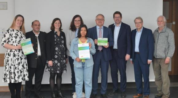 Staatliche Berufsschule Mindelheim Eine Gruppe von neun Personen steht in einem Raum und hält Zertifikate in den Händen. In der Mitte präsentieren zwei Personen stolz ihre Urkunden. Die Gruppe ist gut gekleidet, und die Stimmung ist feierlich. Der Hintergrund ist schlicht und neutral.