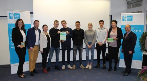 Für das besondere Zusatzangebot und ihre Selbstverpflichtung zu den Zielen des Globalen Lernens erhält die Richard-Müller-Schule Fulda (RiMS) vom World University Service (WUS) die Auszeichnung zur „Grenzenlos-Schule“. Das Bild zeigt eine Gruppe von neun Personen, die in einem Raum zusammenstehen und in die Kamera schauen. Einige halten Zertifikate oder Urkunden in den Händen, die sie stolz präsentieren. Die Gruppe besteht aus Lehrkräften und jungen Menschen, die anscheinend eine Auszeichnung erhalten haben. Der Hintergrund ist schlicht mit Bannern, die das Logo einer Schule zeigen. Alle Teilnehmer wirken freundlich und engagiert, und es scheint sich um eine offizielle Zertifizierungszeremonie zu handeln.