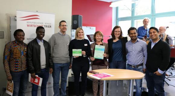 Das Bild zeigt eine Gruppe von neun Personen, die zusammen in einem Raum stehen. Einige von ihnen halten Zertifikate in den Händen und präsentieren diese stolz. Der Hintergrund zeigt ein Banner mit der Aufschrift „Berufliche Schule Wirtschaft Trier“. Es handelt sich vermutlich um eine Auszeichnungsveranstaltung oder ein Event. Der Raum ist gut beleuchtet, mit großen Fenstern und modernen Möbeln. Alle wirken freundlich und engagiert.