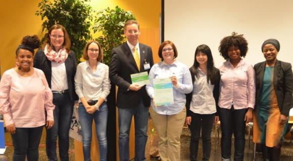 Das Bild zeigt eine Gruppe von sieben Personen, die vor einer Wand mit Pflanzen und einem Präsentationsbild stehen. Einige halten Zertifikate oder Urkunden in den Händen und präsentieren diese stolz. Die Gruppe besteht aus Frauen und einem Mann, die in formeller oder legerer Kleidung gekleidet sind. Alle blicken freundlich in die Kamera. Der Hintergrund ist hell und freundlich, was auf eine feierliche Auszeichnungsveranstaltung hindeutet.