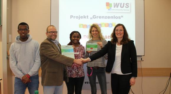 Die Berufliche Schulen am Gradierwerk (BSG) wird „WUS-Grenzenlos-Schule“ und ist regionales Vorbild für mit Freude gelebte Vielfalt und Internationalität. Das Bild zeigt eine feierliche Szene, bei der vier Personen sich die Hände schütteln. Zwei von ihnen halten Zertifikate in der Hand. Es handelt sich um eine Übergabe im Rahmen des Projekts „Grenzenlos“, wie das Plakat im Hintergrund zeigt. Die Personen auf dem Bild stehen nebeneinander: Ein Mann und eine Frau in formeller Kleidung sowie zwei junge Menschen in lockerer Kleidung. Der Raum wirkt modern und gut beleuchtet, und es scheint sich um eine Zeremonie oder ein offizielles Treffen zu handeln.