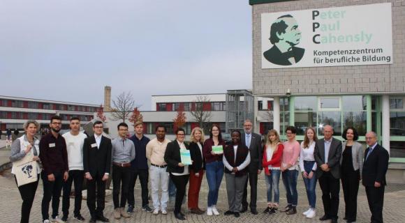 Das Bild zeigt eine Gruppe von 16 Personen vor einem Gebäude mit dem Logo „Peter-Paul-Cahensly Kompetenzzentrum für berufliche Bildung“. Die Gruppe besteht aus Jugendlichen, Erwachsenen und Lehrkräften. Einige Personen halten Zertifikate in den Händen. Alle schauen freundlich in die Kamera. Im Hintergrund ist das moderne Gebäude mit großen Fenstern zu sehen. Es handelt sich vermutlich um eine Zeremonie oder eine Veranstaltung zur beruflichen Weiterbildung.