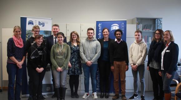 Die Beruflichen Schulen in Groß Gerau sind „WUS-Grenzenlos-Schule“ und kooperieren mit regionalen und internationalen Partnerorganisationen. Das Bild zeigt eine Gruppe von 12 Personen, die in einem Raum zusammenstehen. Sie sind zwischen 15 und 30 Jahre alt und tragen verschiedene Alltagskleidung. Die meisten stehen nebeneinander, einige sind leicht nach vorne gebeugt. Im Hintergrund befinden sich Regale und zwei Banner mit dem Logo "BGS". Es herrscht eine freundliche Atmosphäre, und alle blicken in die Kamera. Einige Personen tragen Brillen oder haben unterschiedliche Frisuren. Es scheint sich um ein Treffen oder eine Veranstaltung zu handeln.