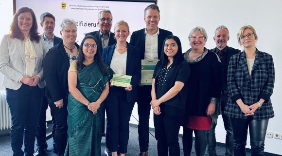 Seminar für Ausbildung und Fortbildung der Lehrkräfte Eine Gruppe von 13 Personen steht in einem hellen Raum vor einer Präsentationsfolie. Zwei Personen in der Mitte halten Zertifikate in den Händen. Die Gruppe lächelt in die Kamera, die Stimmung ist professionell und feierlich. Im Hintergrund ist das Logo des Kultusministeriums Baden-Württemberg zu erkennen.