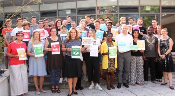 Das Bild zeigt eine große Gruppe von etwa 30 Personen, die vor einem Gebäude im Freien stehen. Alle halten Plakate in den Händen, die verschiedene Ziele der nachhaltigen Entwicklung (SDGs) zeigen. Die Gruppe setzt sich aus Jugendlichen und Erwachsenen unterschiedlicher Herkunft und Geschlechter zusammen. Sie tragen sommerliche Kleidung und wirken stolz und engagiert. Der Hintergrund zeigt große Fenster des Gebäudes und einige Bäume, was der Szene eine freundliche Atmosphäre verleiht.
