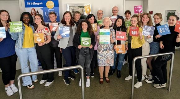 inlingua Augsburg Eine Gruppe von etwa 20 Personen steht in einem Innenraum und hält bunte Schilder mit Symbolen der UN-Nachhaltigkeitsziele (SDGs) hoch. Eine Person in der Mitte präsentiert ein Zertifikat. Im Hintergrund sind Roll-Ups mit Logos und weiteren Informationen zu sehen. Die Stimmung ist fröhlich und engagiert.