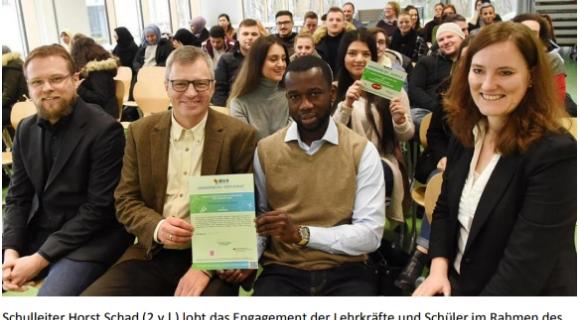 Auf dem Bild sind drei Personen im Vordergrund zu sehen, die jeweils ein Zertifikat in den Händen halten. In der Mitte sitzt ein Mann, der das Zertifikat zusammen mit einer weiteren Person hält. Links und rechts von ihnen sitzen eine weitere Person und eine Frau. Im Hintergrund ist eine Gruppe von Menschen in einem Veranstaltungsraum zu sehen. Das Bild zeigt eine feierliche Szene, bei der das Engagement von Lehrkräften und Schülern gewürdigt wird.